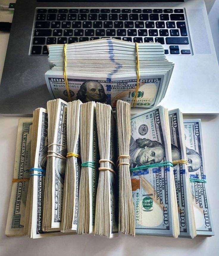 stacks of money sitting in front of a laptop on top of a white desk next to a pile of hundred dollar bills