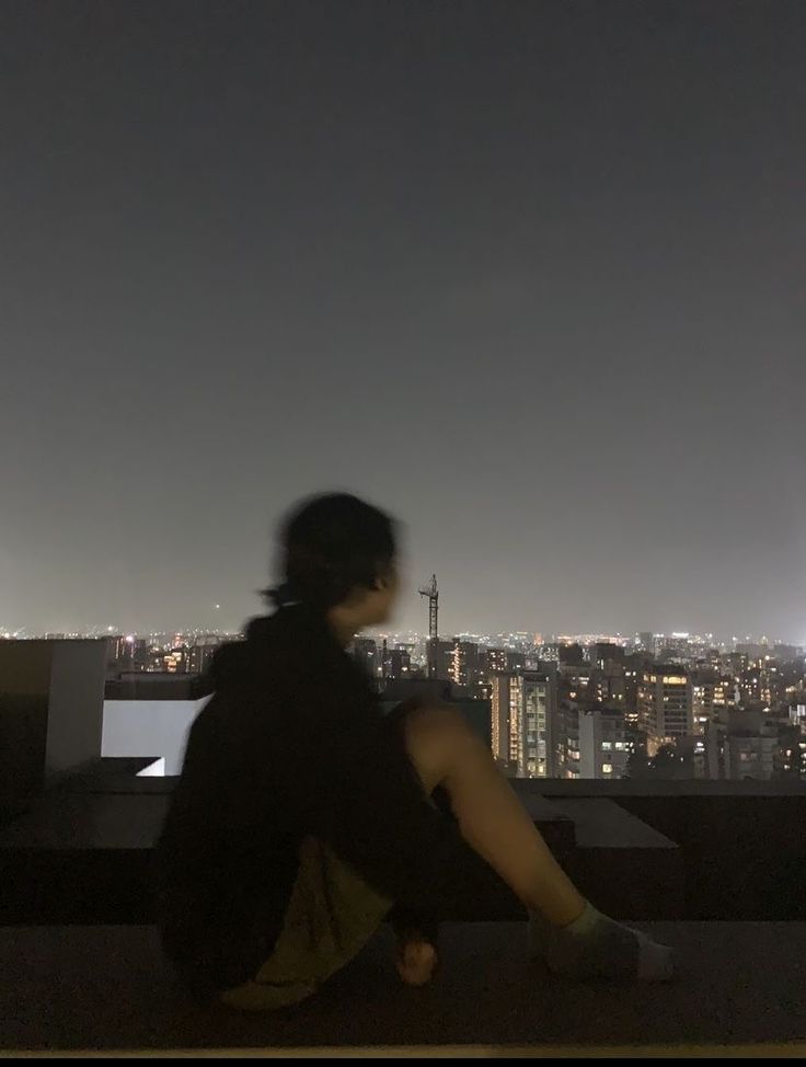 a woman sitting on top of a roof looking at the city lights in the distance