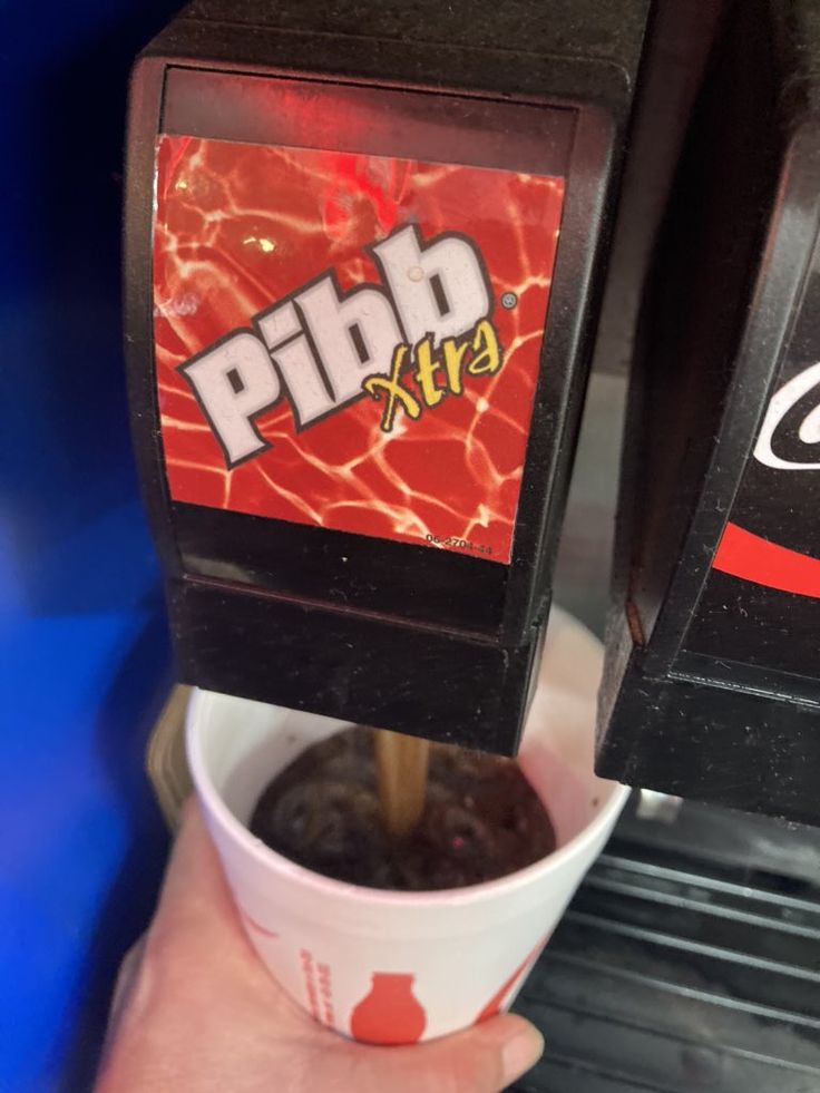 a person is holding a cup in front of a machine that sells chips and drinks