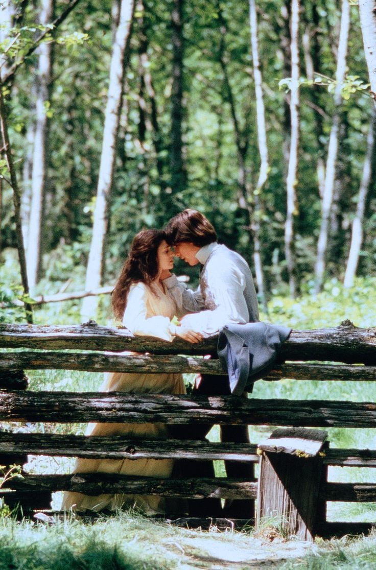 a man and woman sitting on a bench in the woods, looking at each other