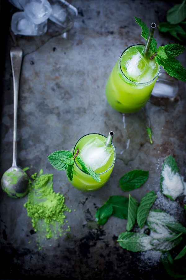 two glasses filled with green drinks next to mint leaves and spoons on a table