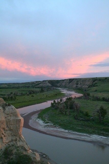 a river running through a lush green field next to a rocky hillside under a pink sky