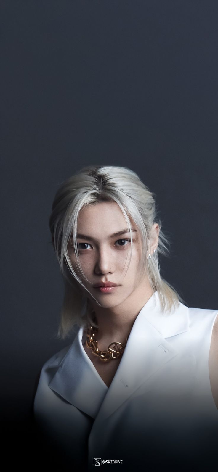 a woman with blonde hair wearing a white shirt and gold necklace, looking at the camera