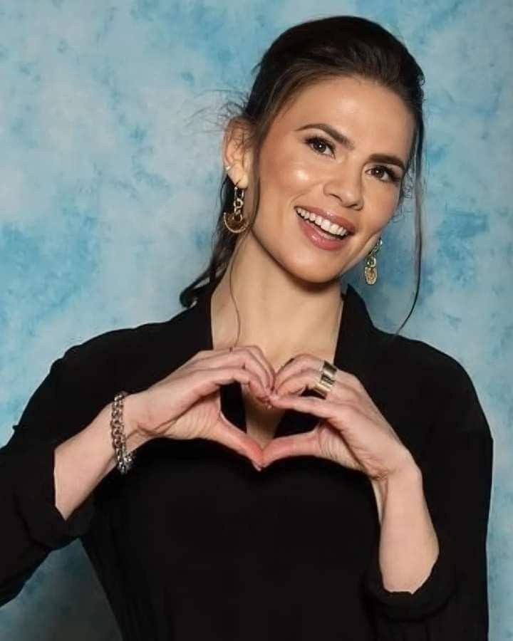 a woman making a heart shape with her hands and smiling at the camera while standing in front of a blue background