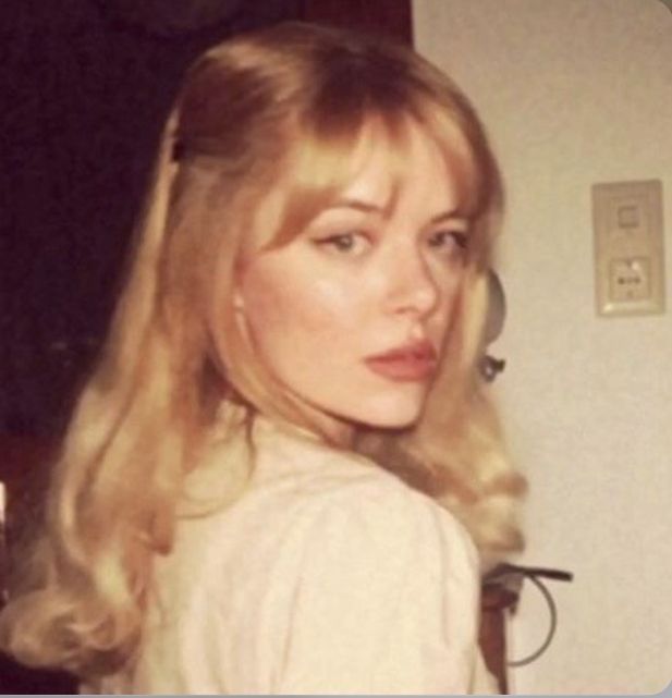 a woman with long blonde hair standing in front of a wall and looking at the camera