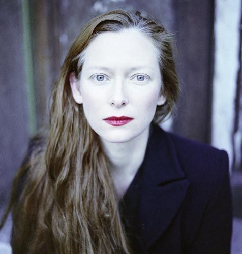 a woman with long hair wearing a black jacket and red lipstick looking at the camera