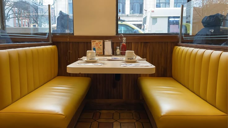 a booth with two yellow benches and a white table in front of large windows that look out onto the street