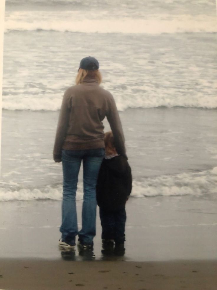 a woman and child standing on the beach looking out at the waves in the ocean