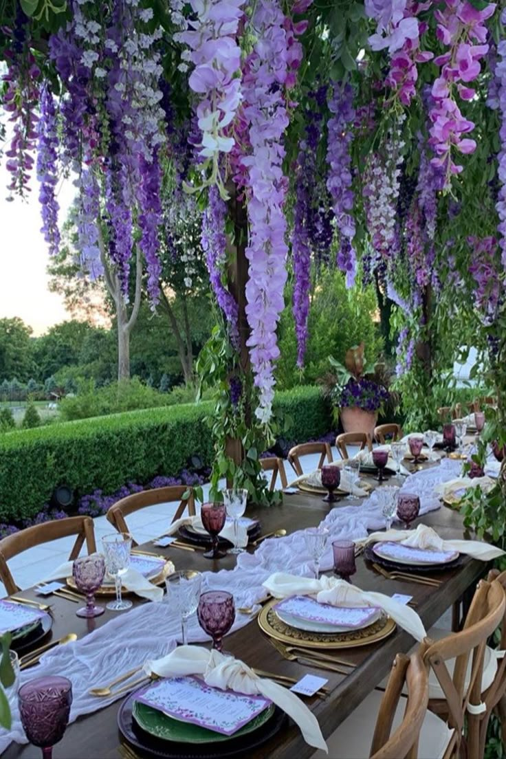 an outdoor dining area with purple flowers hanging from the ceiling and place settings on wooden chairs