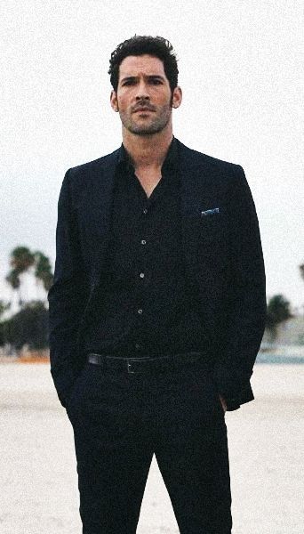 a man in a black suit standing on the beach with his hands in his pockets