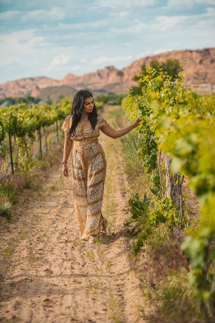 a woman walking down a dirt road next to a lush green field with lots of plants