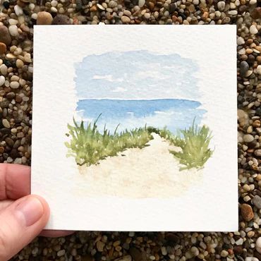 a hand holding up a watercolor painting of a path to the ocean with grass and rocks