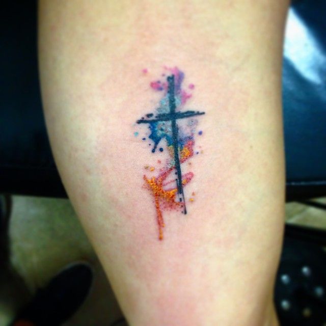 a cross tattoo on someone's leg with watercolor paint splatters all over it