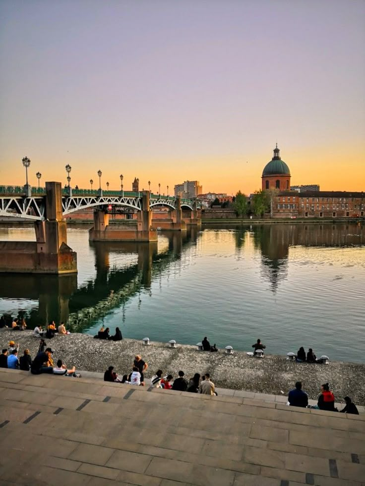 people sitting on the edge of a river watching the sun set