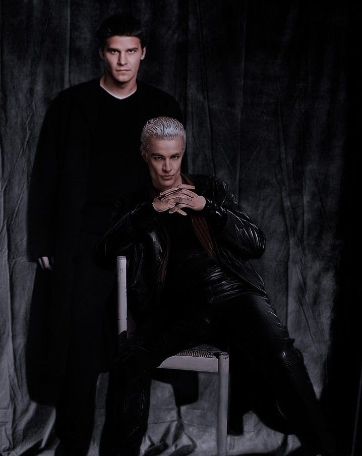 two men are posing for a photo in front of a dark background, one is sitting on a chair and the other is standing