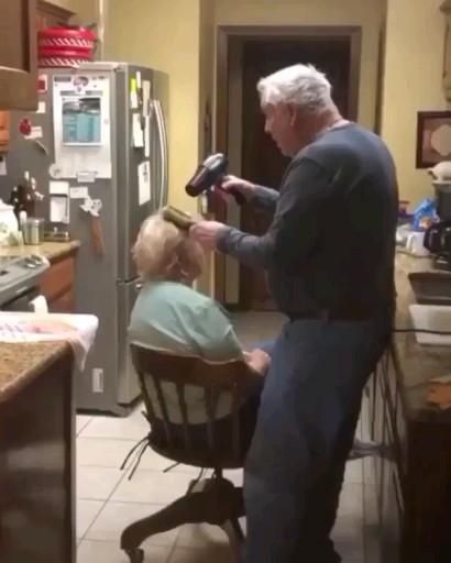 an older man sitting in a chair with a young boy on his lap while he is using a hair dryer