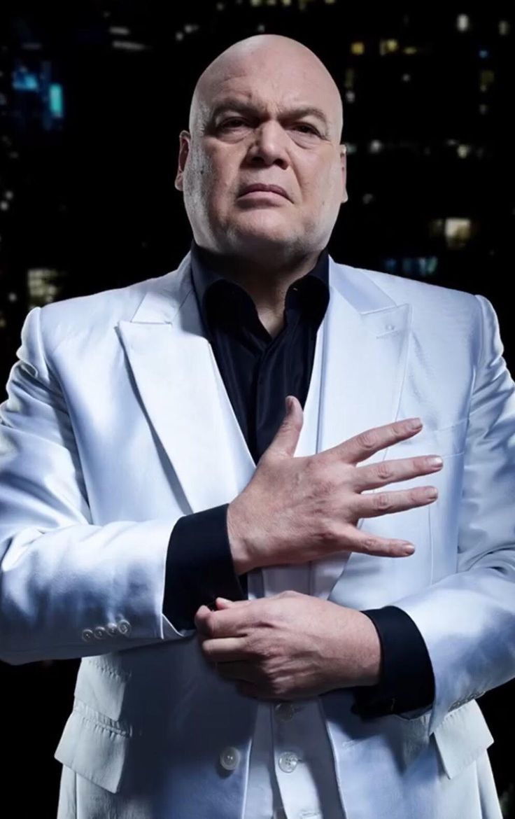 a bald man wearing a white suit and black shirt with his hands folded out in front of him