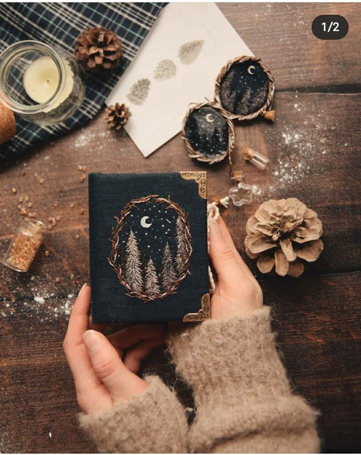 a person holding a small book on top of a wooden table next to pine cones