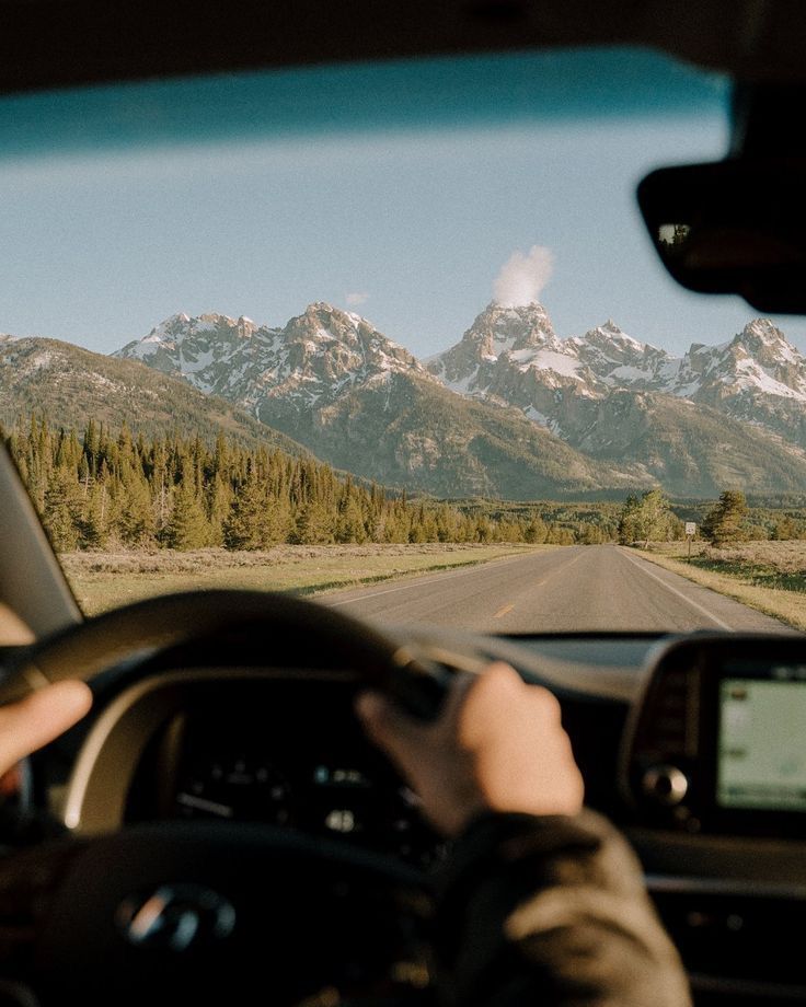 a person driving a car on a road with mountains in the background and snow capped peaks