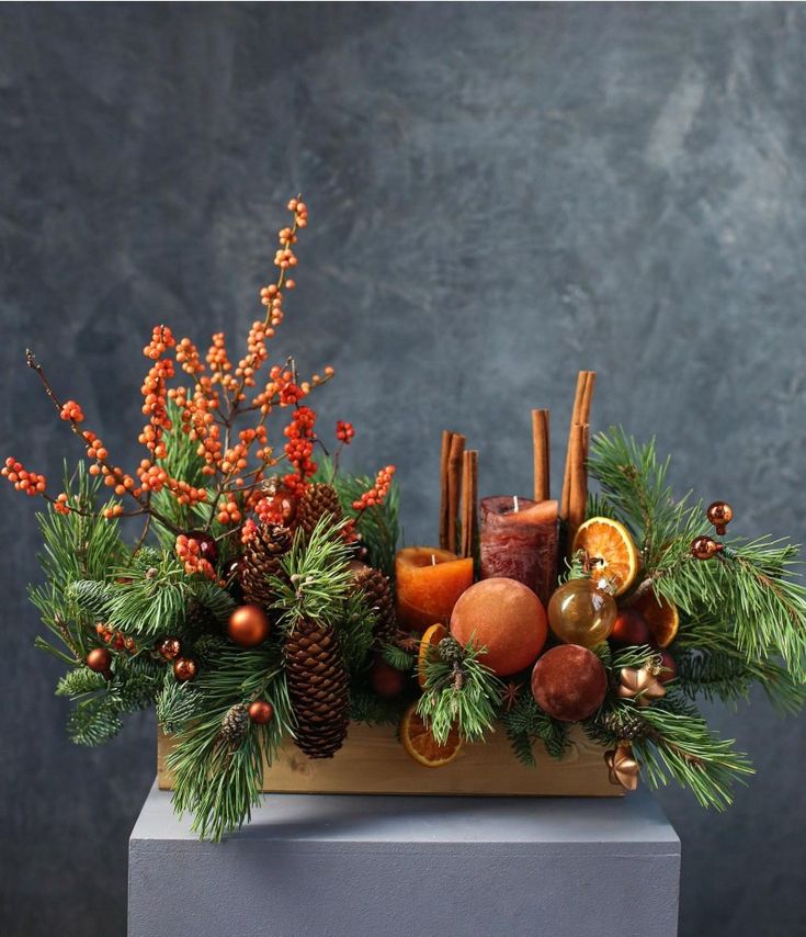 an arrangement of fruit and greenery is displayed on a block with pine cones, berries, cinnamon sticks, oranges, and other holiday decorations