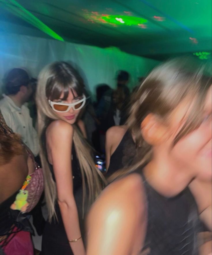 two women in black dresses are dancing at a party with other people and one woman is wearing sunglasses