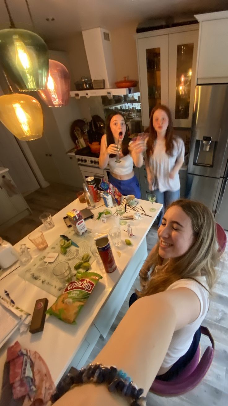 three women standing around a kitchen counter with food on the table and drinks in front of them