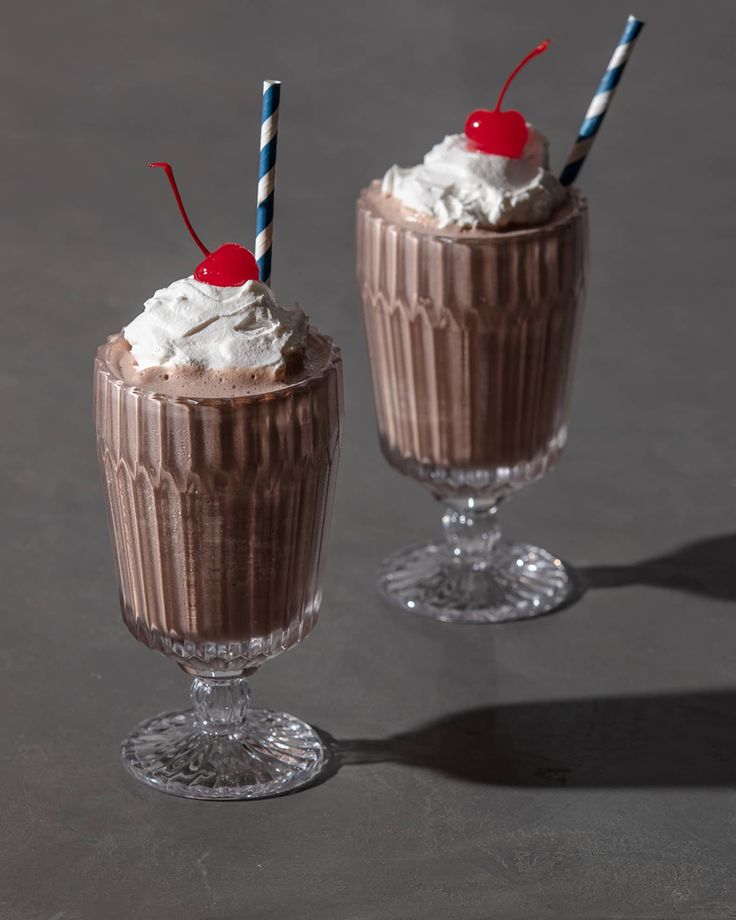 two glasses filled with chocolate pudding and topped with whipped cream, cherries and blue and red striped straws