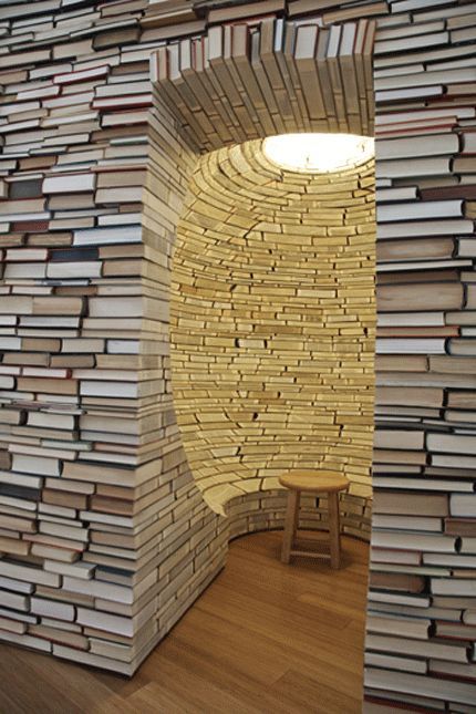 a room made out of books with a lamp on the wall and a stool in front of it