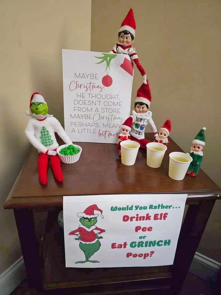 an elf is sitting on top of a table next to cups and a sign that says, would you eat grinch pop?