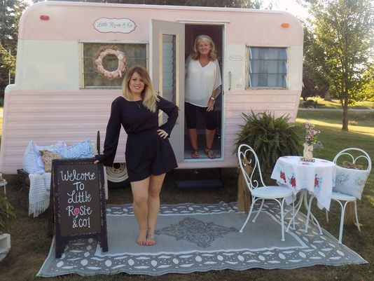 two women standing in the doorway of a pink trailer with a chalkboard sign next to it