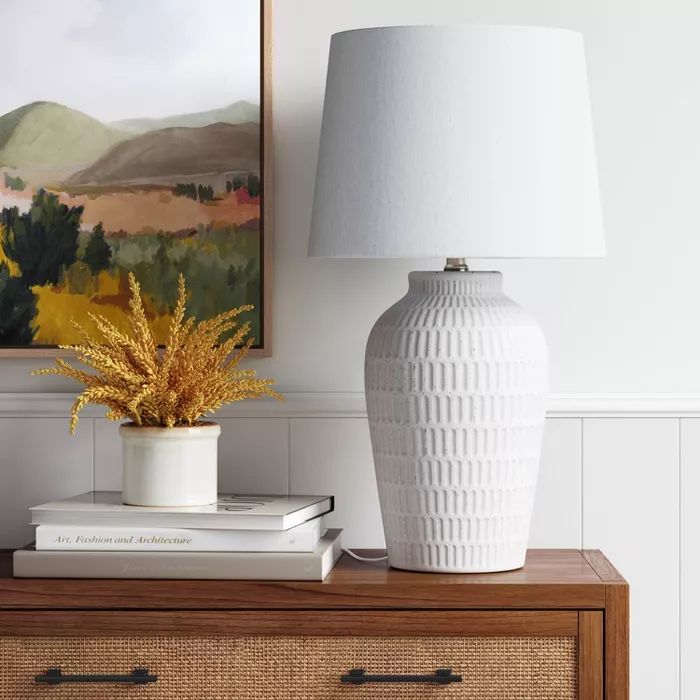 a white vase sitting on top of a wooden table next to a book and lamp