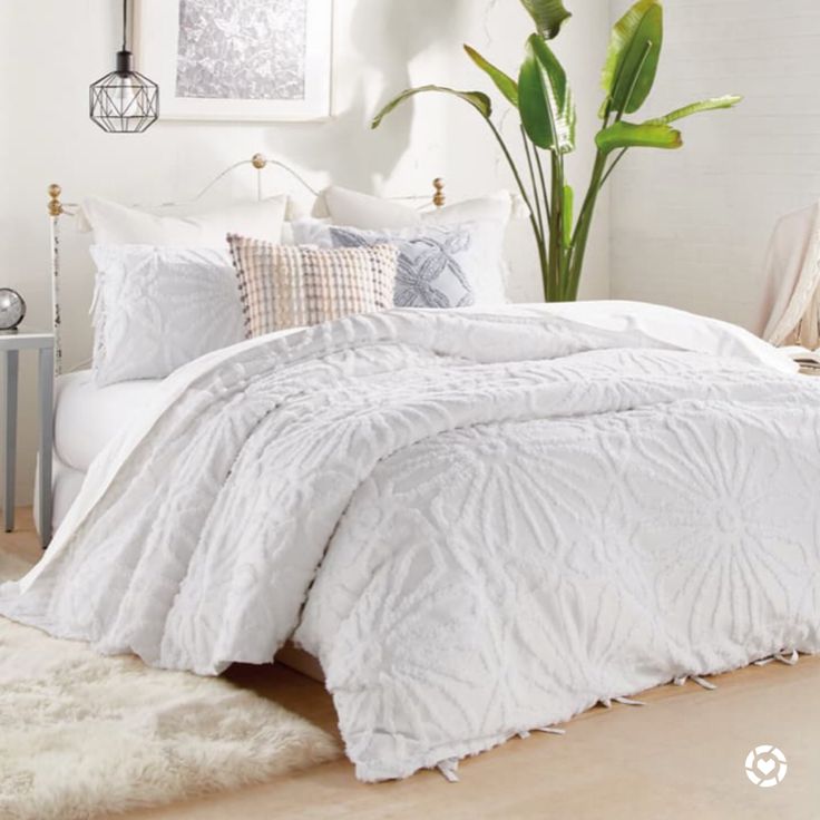 a bed with white comforter and pillows in a room next to a plant on the floor