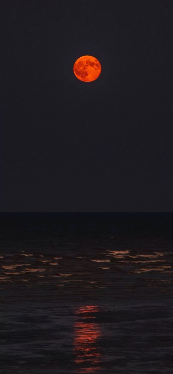 the full moon is seen over the ocean at night with bright orange lights on it