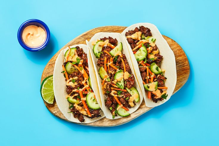 three tacos with meat and vegetables on a wooden platter next to a bowl of ranch dressing