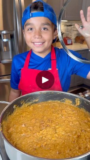 a young boy wearing an orange apron and blue bandana holding up a metal pan with food in it