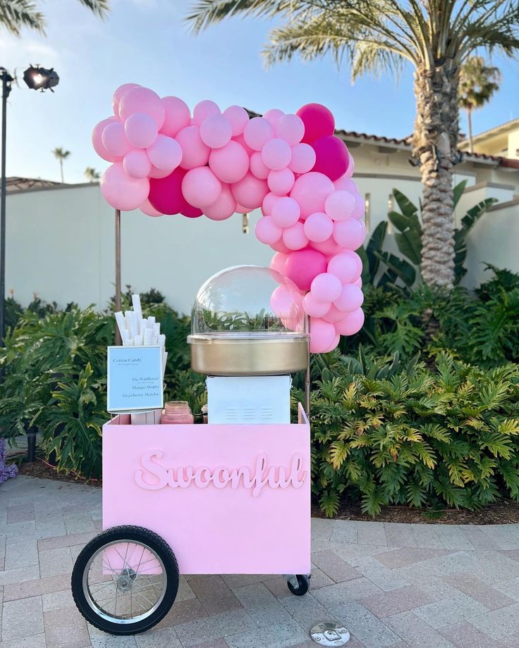 an ice cream cart decorated with pink balloons and writing on the front, in front of a palm tree