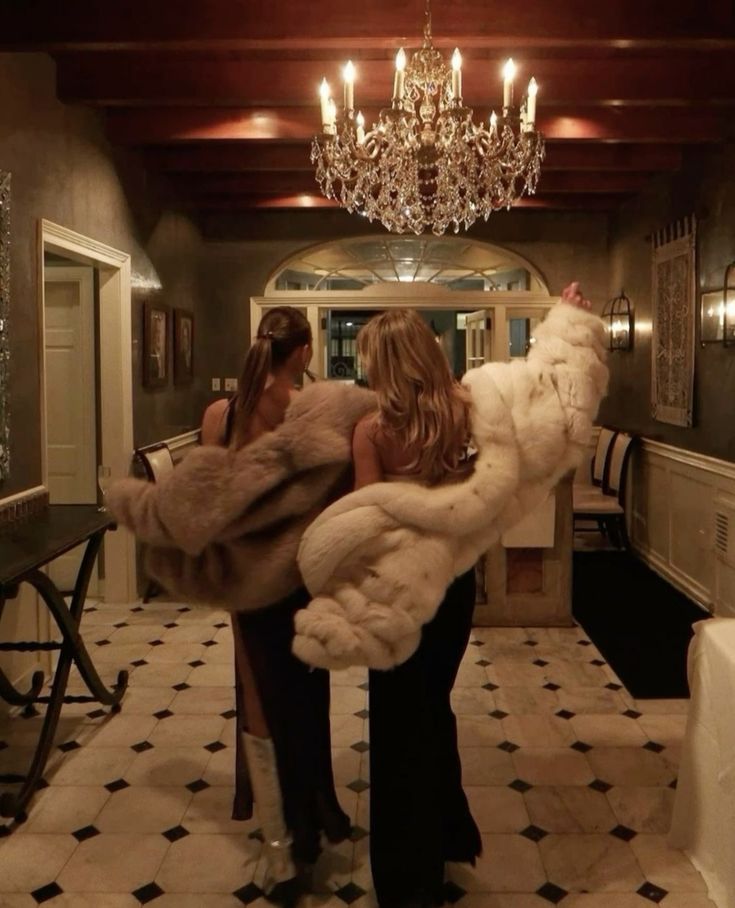 two women standing in a hallway with chandelier and fur coats over their shoulders