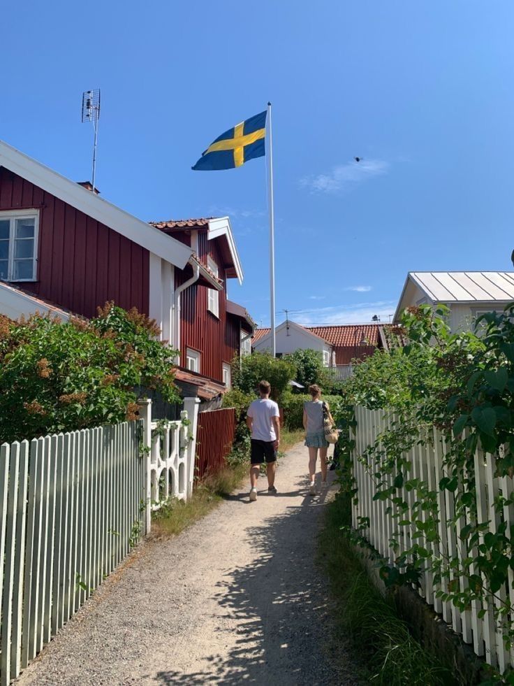 two people walking down a dirt road next to houses and a swedish flag flying in the background