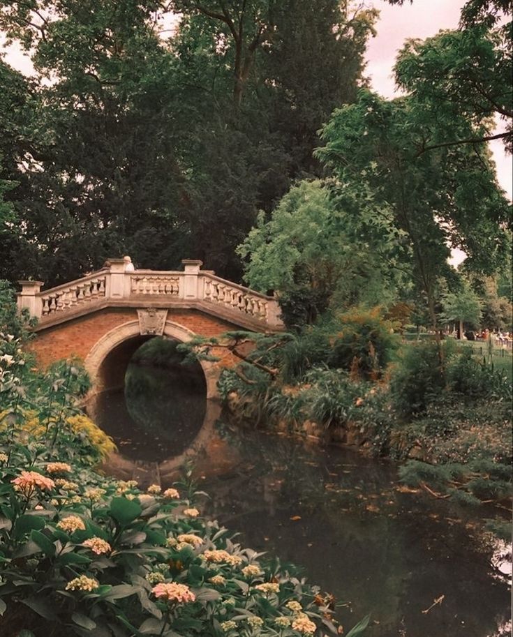 a bridge over a small river surrounded by lush green trees and flowers in the foreground