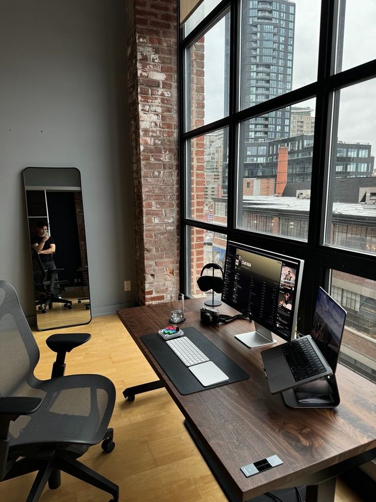 a desk with a laptop, monitor and phone on it in front of large windows