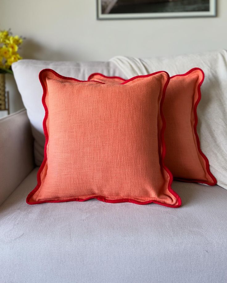 two orange and red pillows sitting on top of a white couch next to a vase with yellow flowers