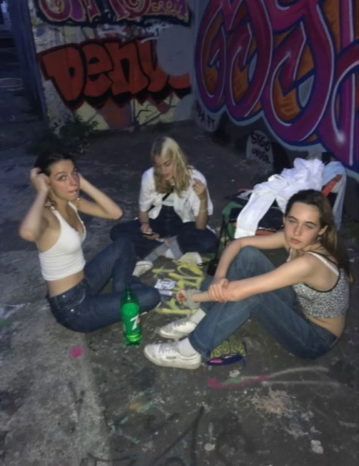 three young women sitting on the ground in front of graffiti covered walls, one is holding her head