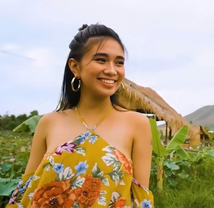 a woman standing in the middle of a field with flowers on her dress and large hoop earrings