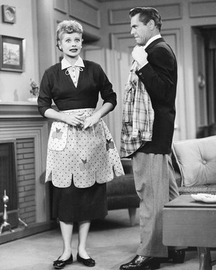 an old photo of two people standing in a living room with fireplace and couches