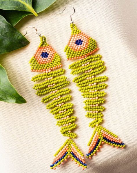 two green and orange beaded fish earrings sitting on top of a white cloth next to a plant