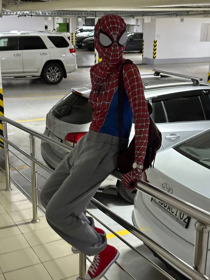 a man in a spiderman costume is sitting on a rail near parked cars at an airport