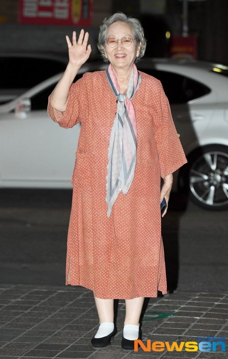 an older woman waves while standing in front of a white car