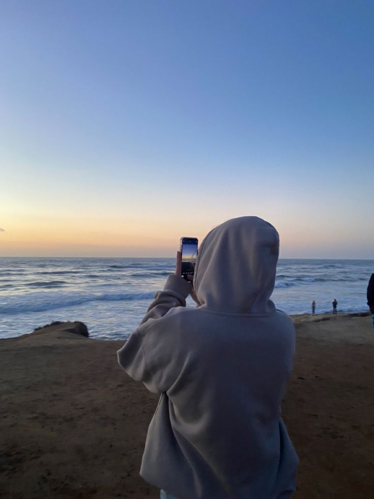 a person wearing a hoodie taking a photo with their cell phone at the beach