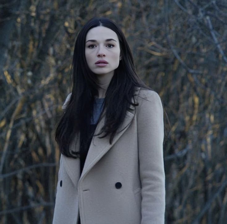 a woman with long black hair standing in front of trees and looking off into the distance