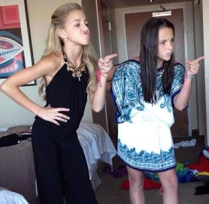 two young women standing in a bedroom pointing at something on the wall behind them,